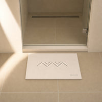 MEGEM Alpine stone bath mat in natural bone on sand tile at the step-out point in front of a glass shower door with a linear drain, soft morning light