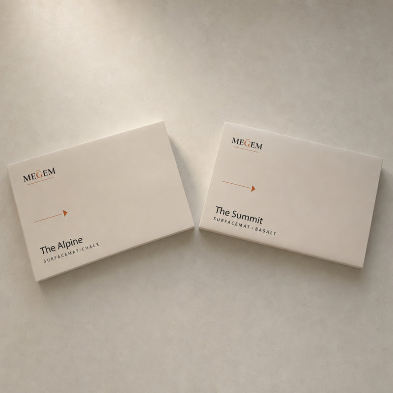 Two closed MEGEM boxes labeled The Alpine and The Summit stone bath mat variants on a pale stone surface