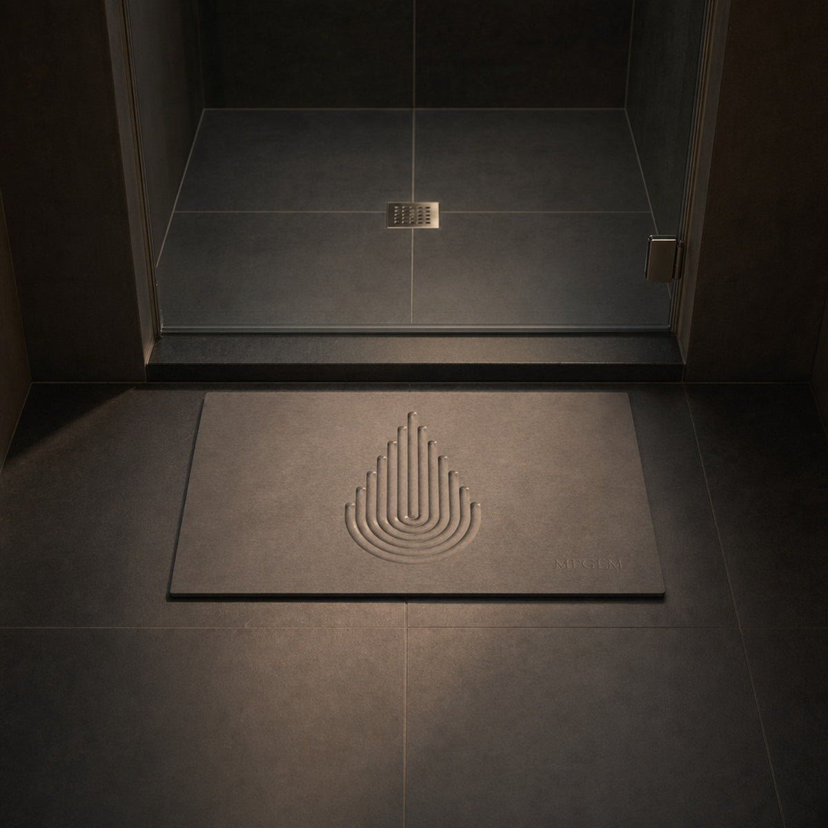 MEGEM Summit stone bath mat in natural grey on charcoal tile in front of a glass shower door with a square drain, low evening light