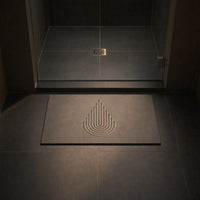 MEGEM Summit stone bath mat in natural grey on charcoal tile in front of a glass shower door with a square drain, low evening light